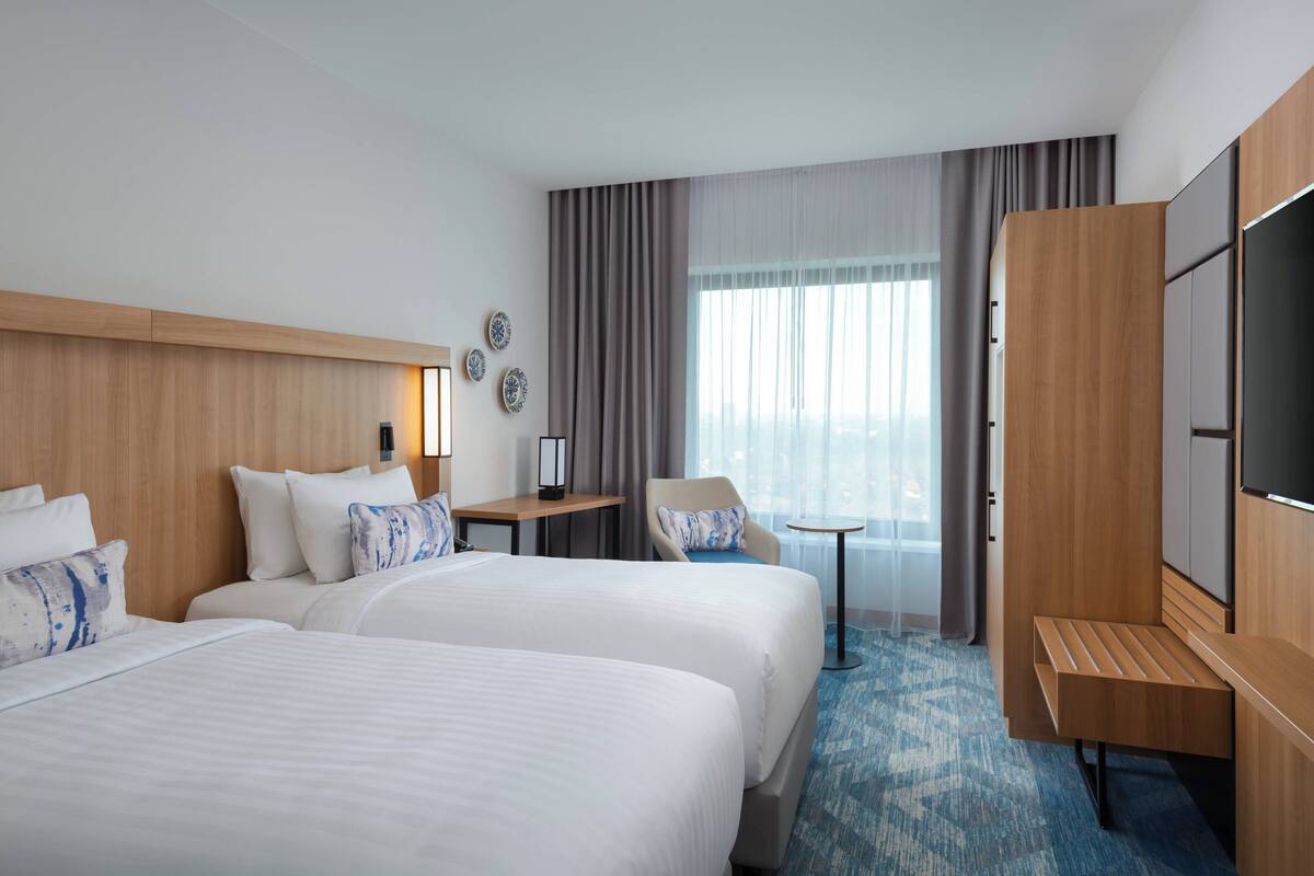 Ein Hotelzimmer mit zwei Betten, einem Schreibtisch, einem Sessel, einem Fernseher und einem großen Fenster mit Vorhängen.