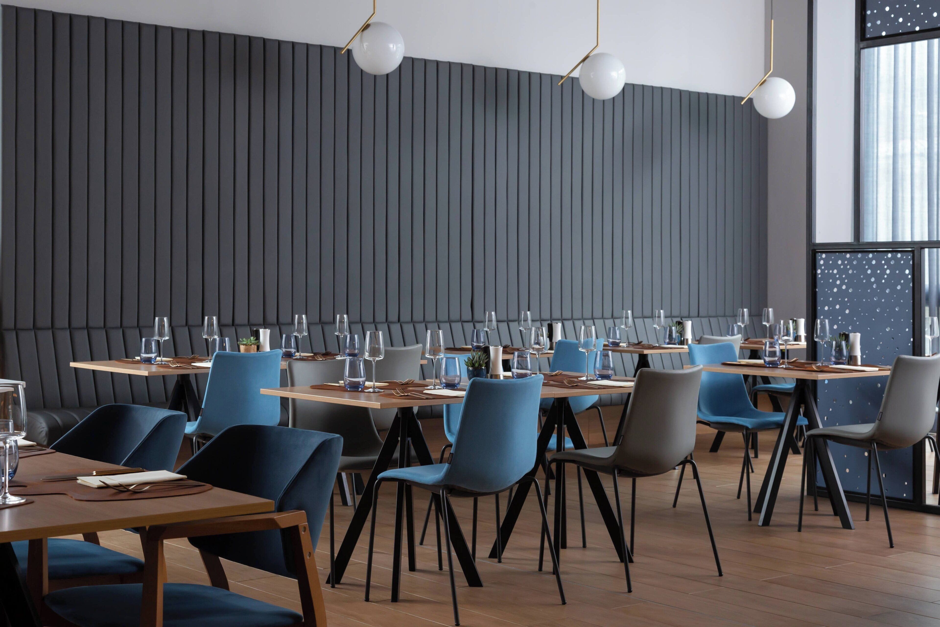A modern dining area with tables set for four, blue and grey chairs, and a dark wooden wall with vertical lines.