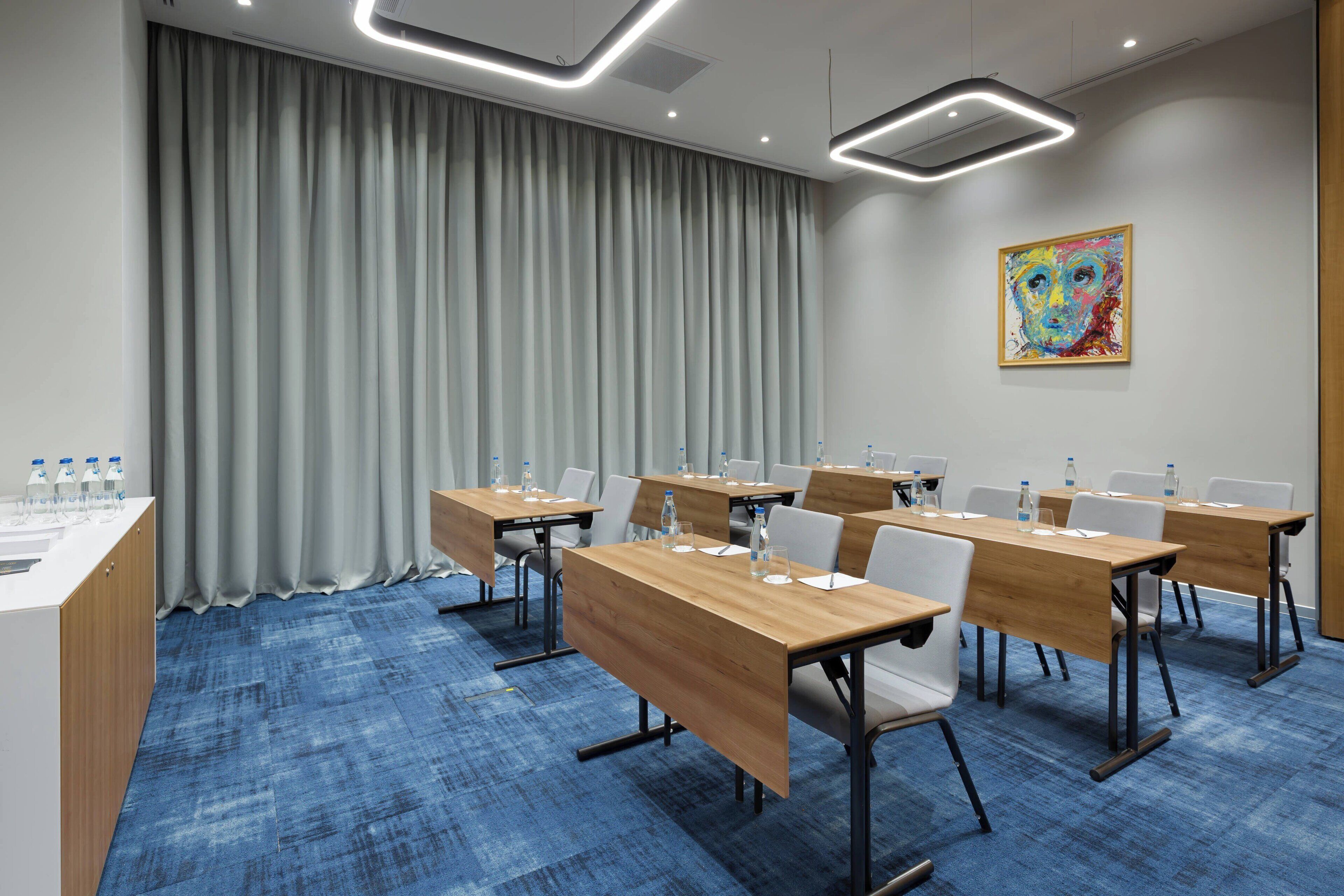 A conference room with a blue carpet, wooden tables, white chairs, a white counter with water bottles, and a colorful painting on the wall.
