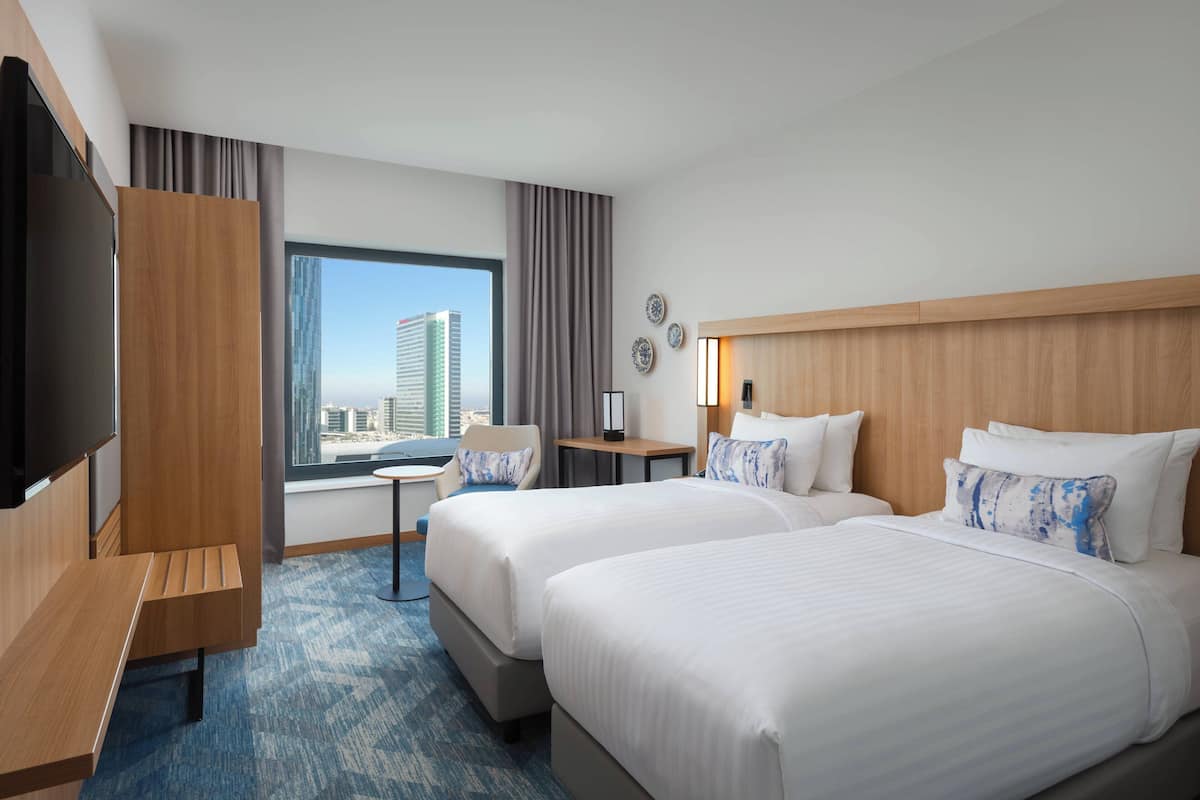 Ein Hotelzimmer mit zwei Betten, einem Schreibtisch, einem Stuhl, einem Fernseher und einem Fenster mit Blick auf die Stadt.