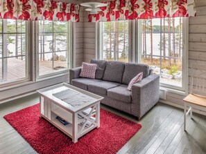 Un salon chaleureux avec un canapé gris, un tapis rouge et de grandes fenêtres donnant sur un paysage naturel.