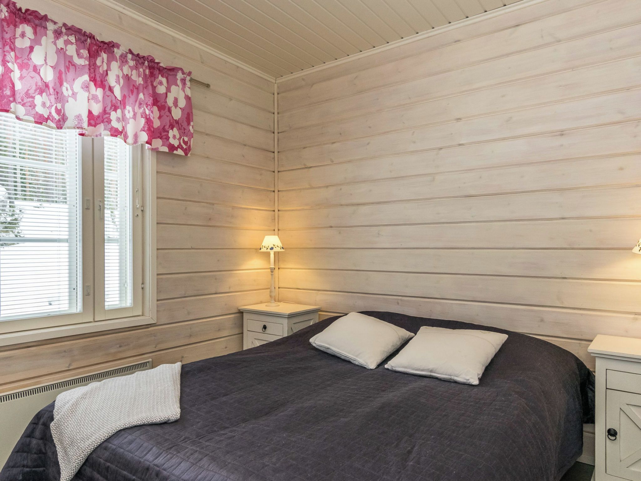 A bedroom with a bed, two bedside tables, a window with blinds, and a wall-mounted lamp.