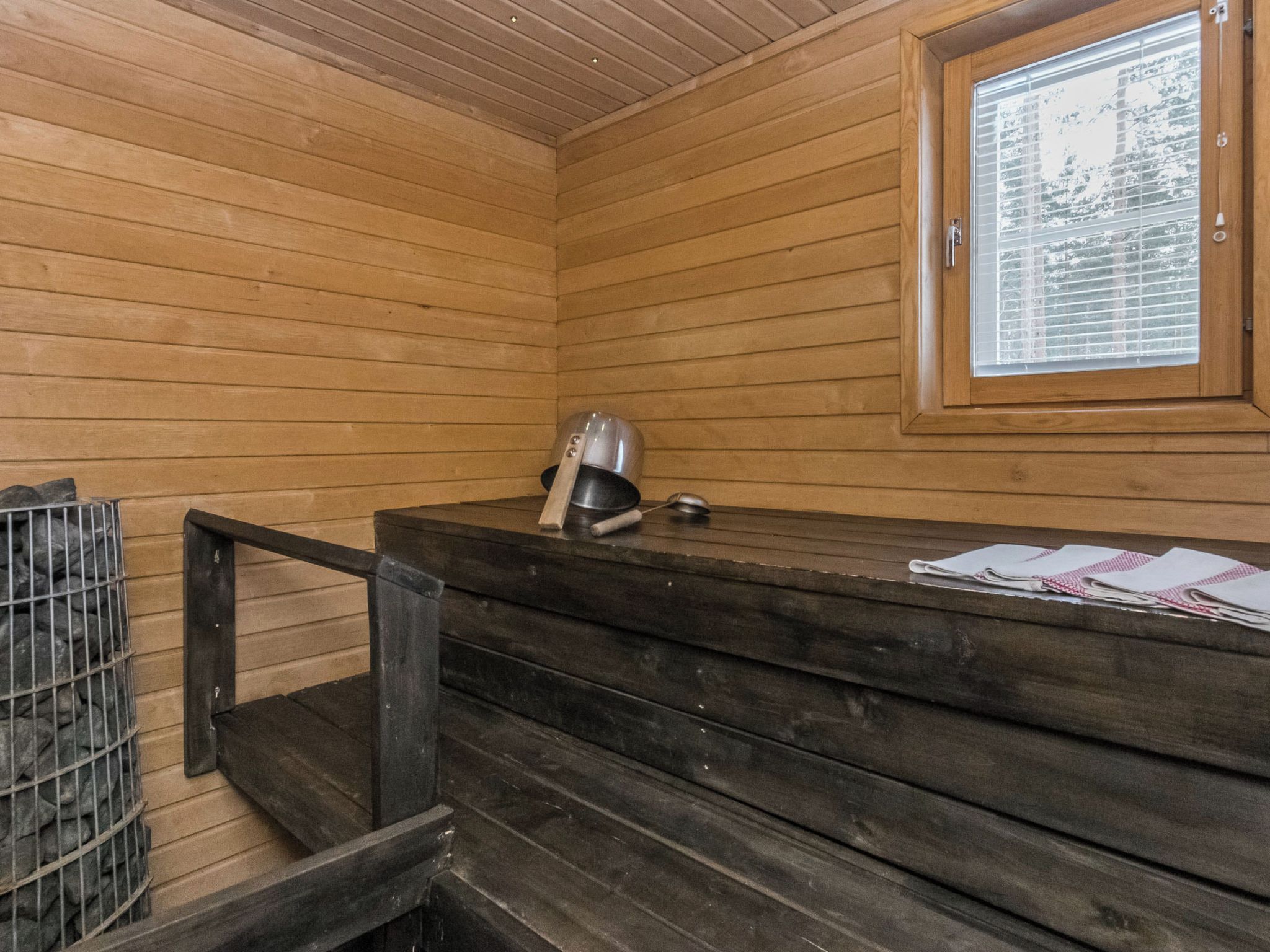 A wooden cabin interior with a bench, a metal grill, and a window with blinds.