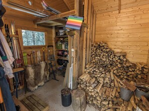 A wooden cabin interior with a ski rack, a pile of firewood, and a window with a snowy view.