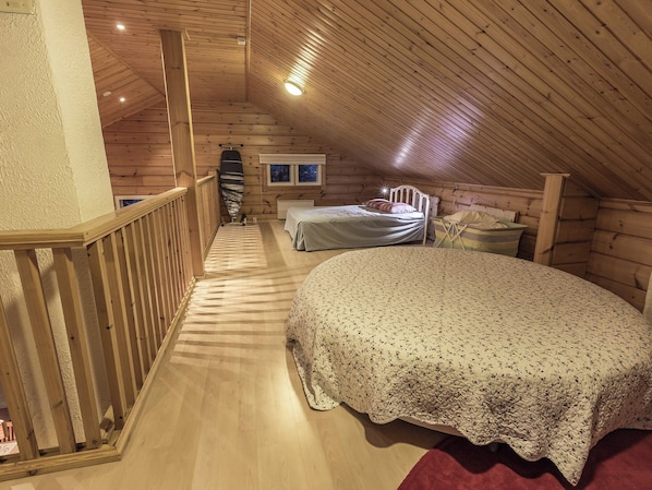 A wooden cabin bedroom with a sloped ceiling, a bed with a floral bedspread, a small table, and a window with a view of the outside.