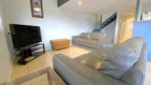 A living room with a flat-screen TV, a grey sofa, a wicker side table, and a staircase leading to an upper floor. - Osprey Holiday Village Unit 122/2 Bedroom apartment in complex with a pool (EXMOUTH)