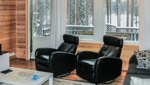 Una stanza accogliente con pareti in legno, un divano in pelle nera e due poltrone in pelle nera. Ampie finestre offrono vista su alberi innevati.