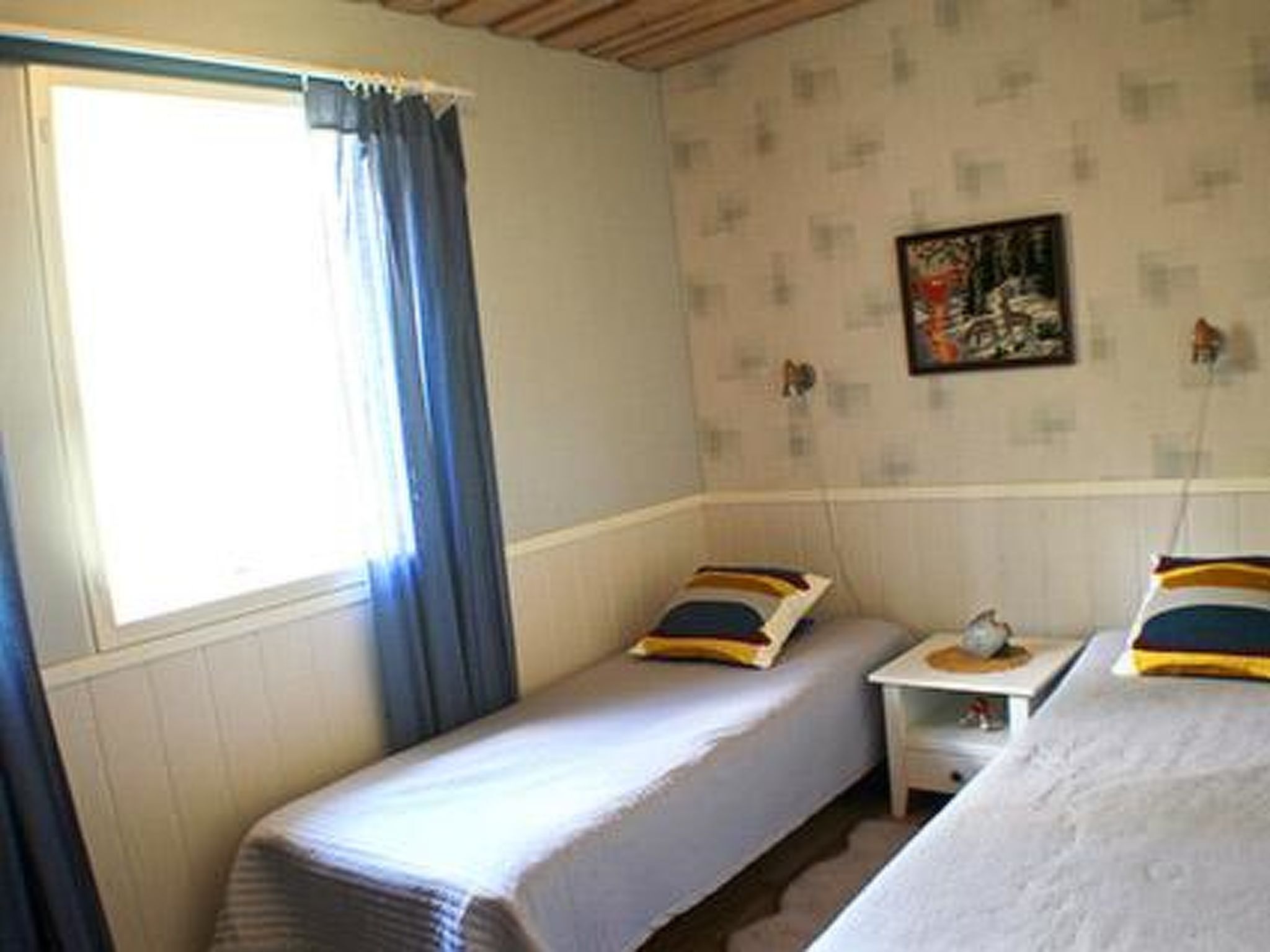 A room with two single beds, a small bedside table with a lamp, a window with curtains, and a wall with a framed picture.