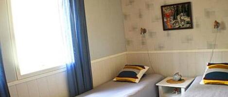 A room with two single beds, a small bedside table with a lamp, a window with curtains, and a wall with a framed picture.