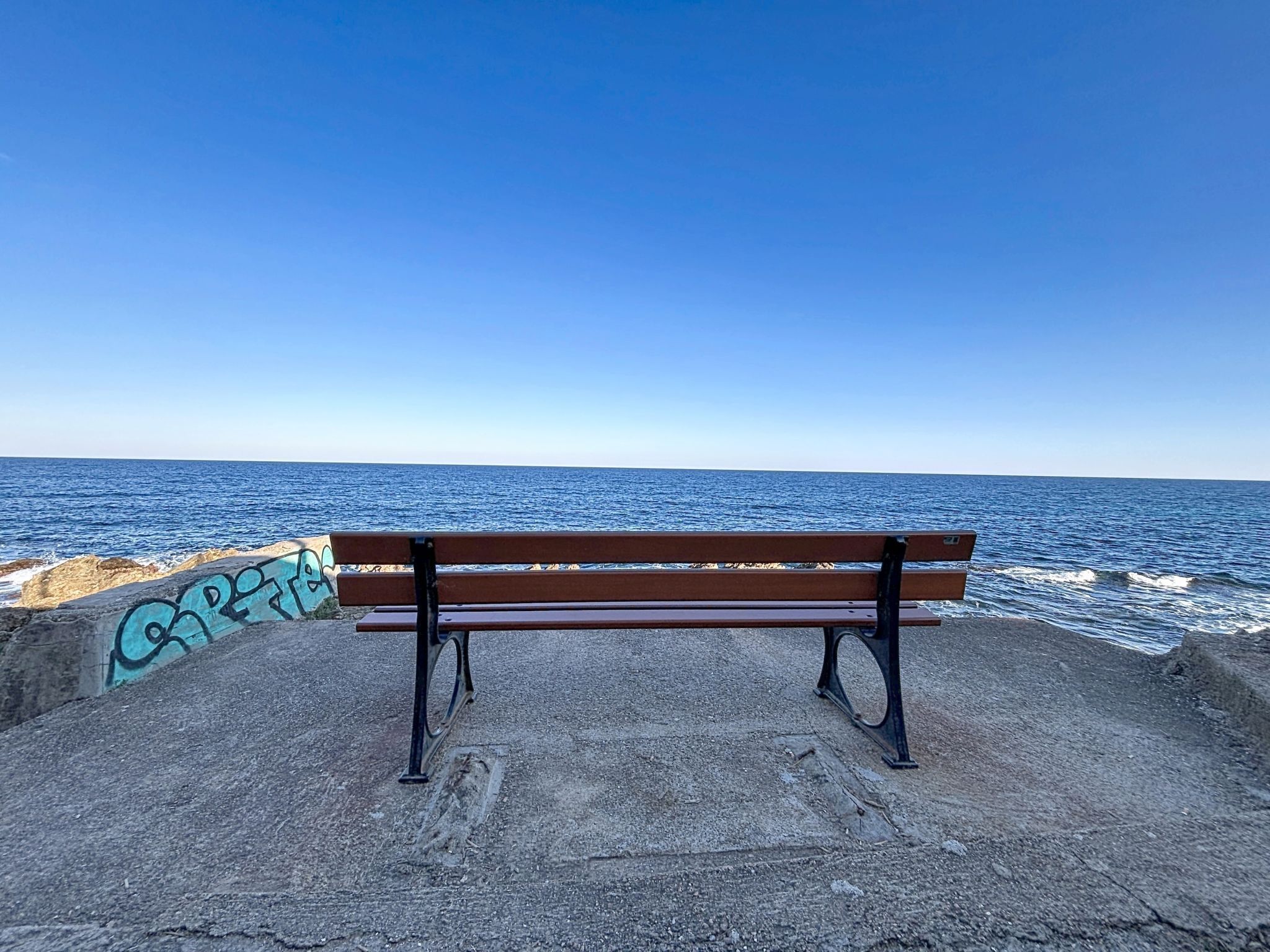A bench by the sea with graffiti on the wall.