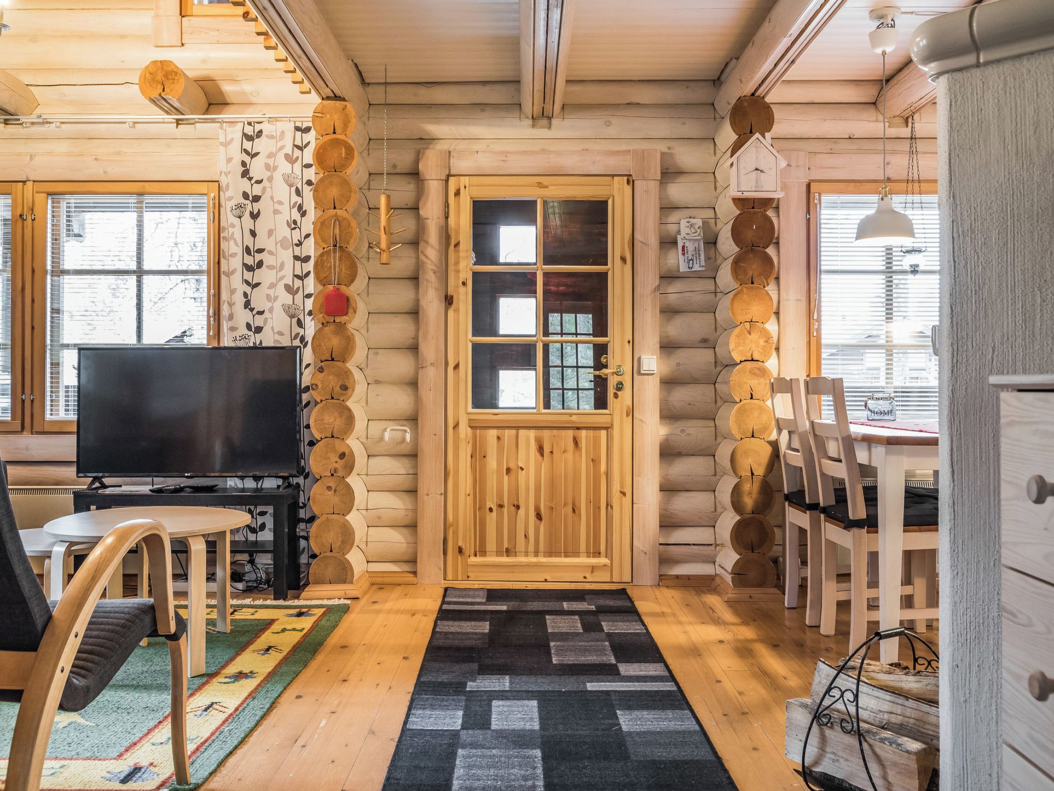 Interior de una cabaña de troncos con televisor, mesa de comedor, sillas y una alfombra estampada.