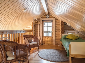A cozy attic bedroom with a wooden sloped ceiling, a bed with green bedding, a wicker chair, and a small rug.