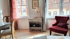Quarto com escrivaninha, cadeira, poltrona de veludo vermelho, radiador e janela com vista para edifícios.