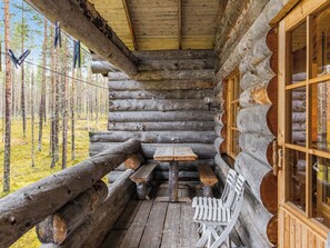 A log cabin with a wooden table and benches, a white chair, and a window with a view of a forest. - Ylläsnäkyy 7 / karkelo by Interhome (Äkäslompolo)