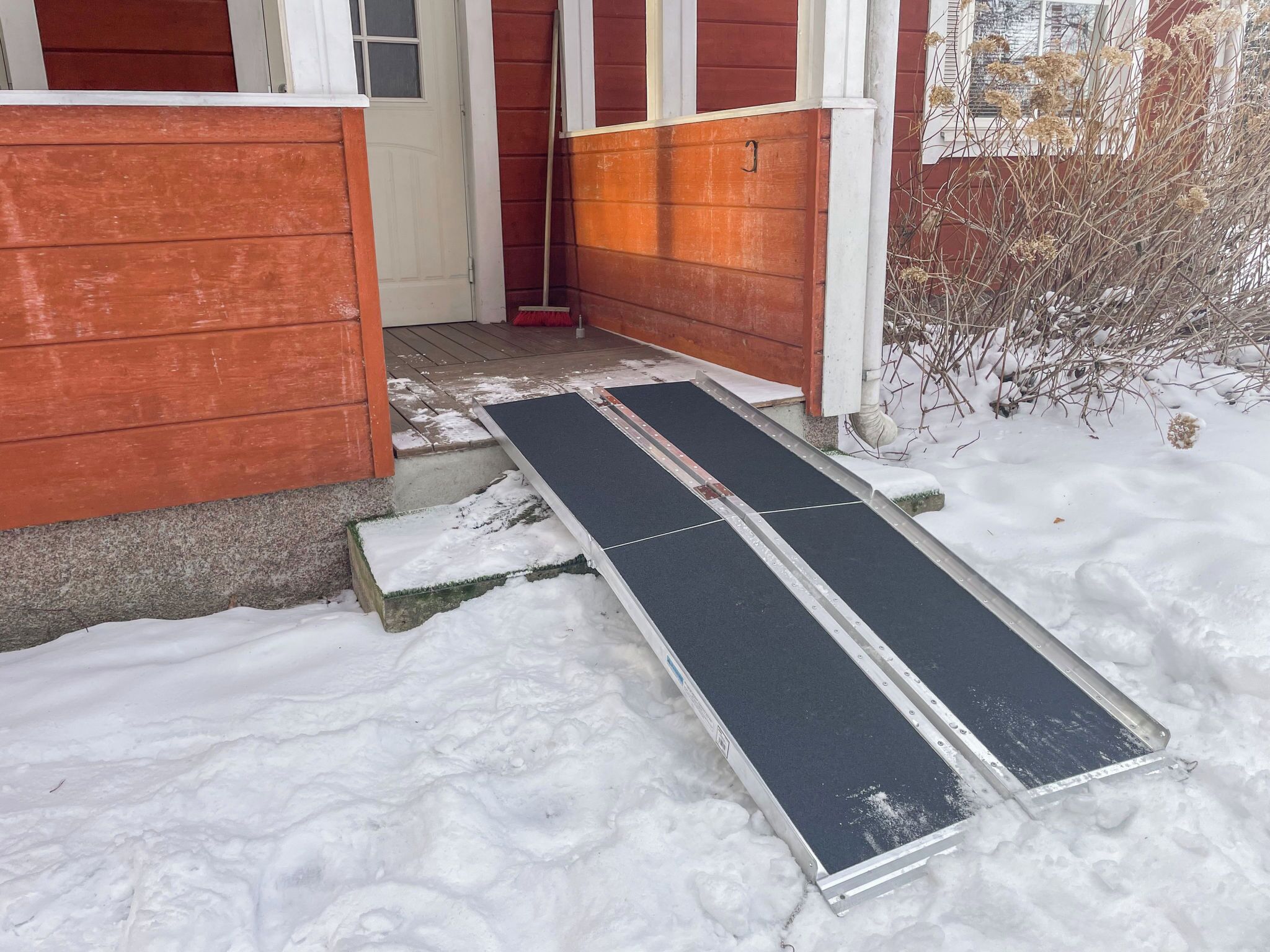 Une maison en bois recouverte de neige, avec une rampe métallique menant à une porte.