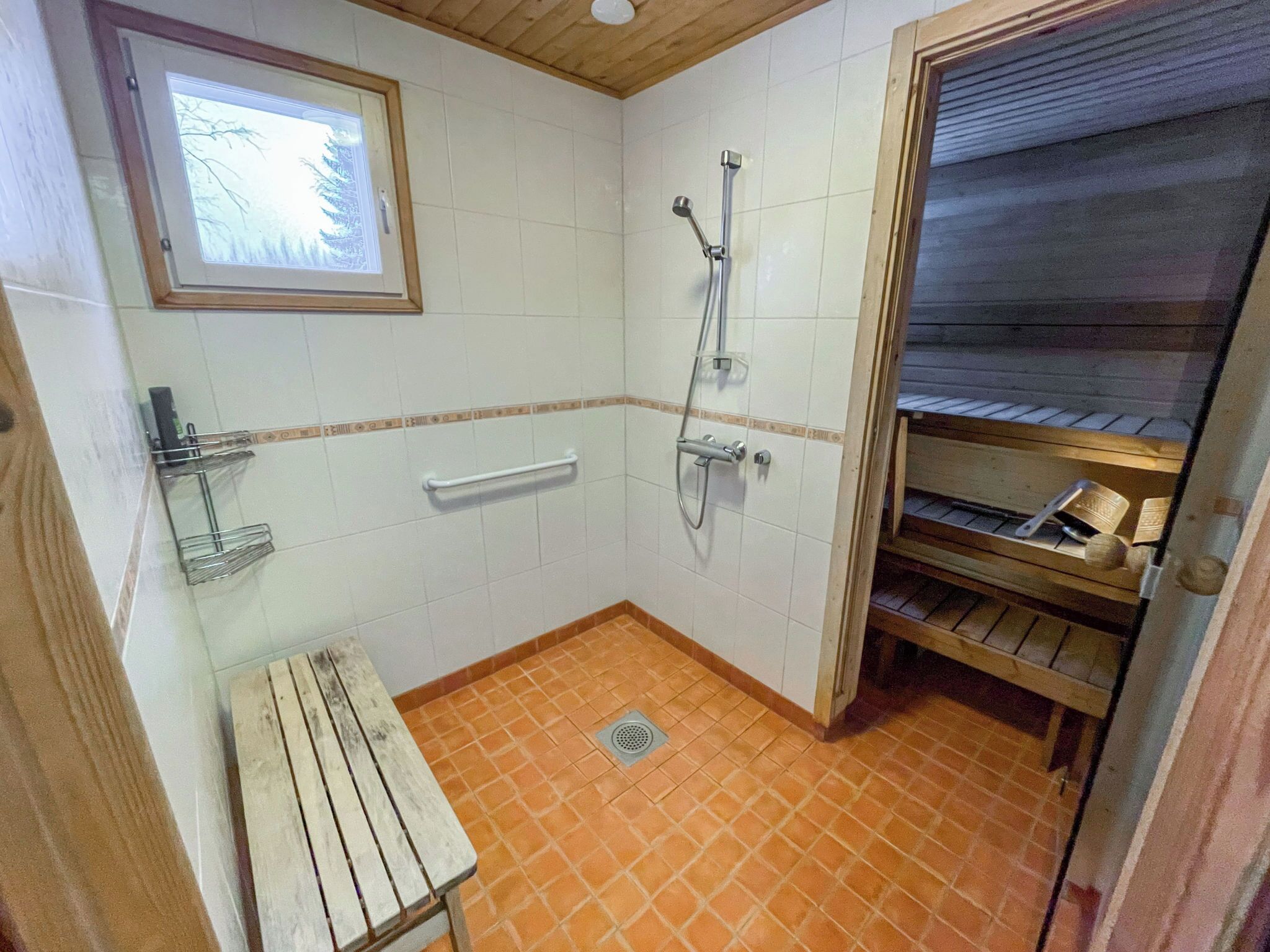 Une petite salle de bain carrelée, équipée d’une douche, d’un banc et d’une étagère où sont rangés les articles de toilette.
