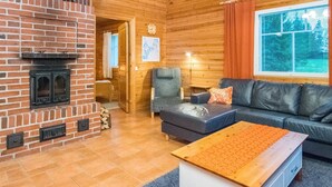 A cozy cabin interior with a brick fireplace, a sofa, a wooden coffee table, and a large window with blinds.