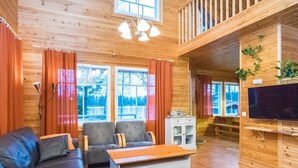 A cozy cabin interior with a sofa, chairs, a small table, a TV, and a balcony with plants.