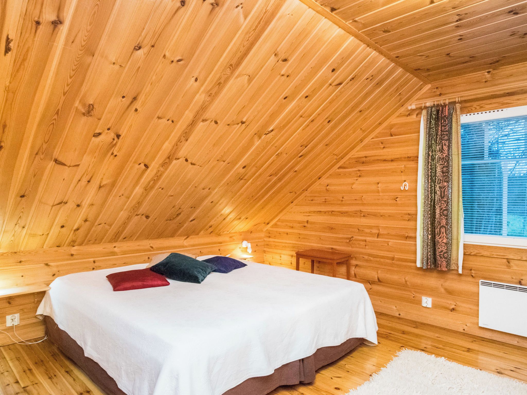 A wooden cabin bedroom with a sloped ceiling, a bed with pillows, a window with curtains, and a white radiator.