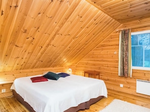 Een slaapkamer in een houten chalet met een schuine kap, een bed met kussens, een raam met gordijnen en een witte radiator.