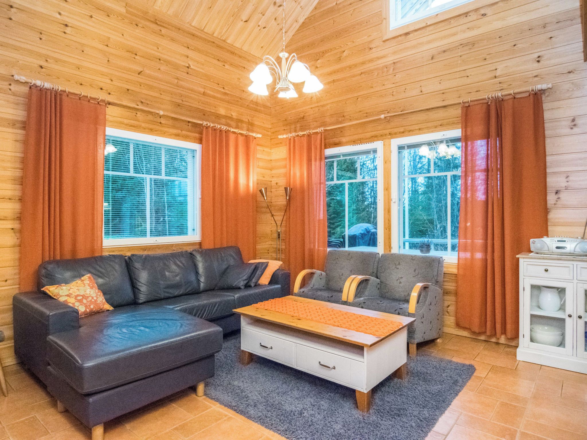 A cozy cabin interior with a sectional sofa, a small table, and two armchairs. There are large windows with curtains, a ceiling fan, and a cabinet with dishes.