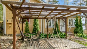 Une terrasse en bois agrémentée d’une pergola, de deux chaises et d’un petit espace jardin avec des plantes en pot et une clôture en bois.