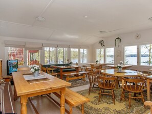 A spacious room with multiple wooden tables and benches, large windows offering views of a lake, and a television on the left.