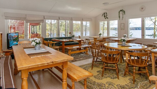 Un vaste espace dédié à la salle à manger, avec des tables et des bancs en bois, de grandes fenêtres offrant une vue sur un lac, et un coin salon confortable avec un canapé et des fauteuils.