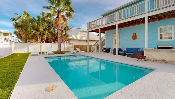 Une piscine avec une terrasse blanche, un patio couvert doté de mobilier d’extérieur et un palmier.