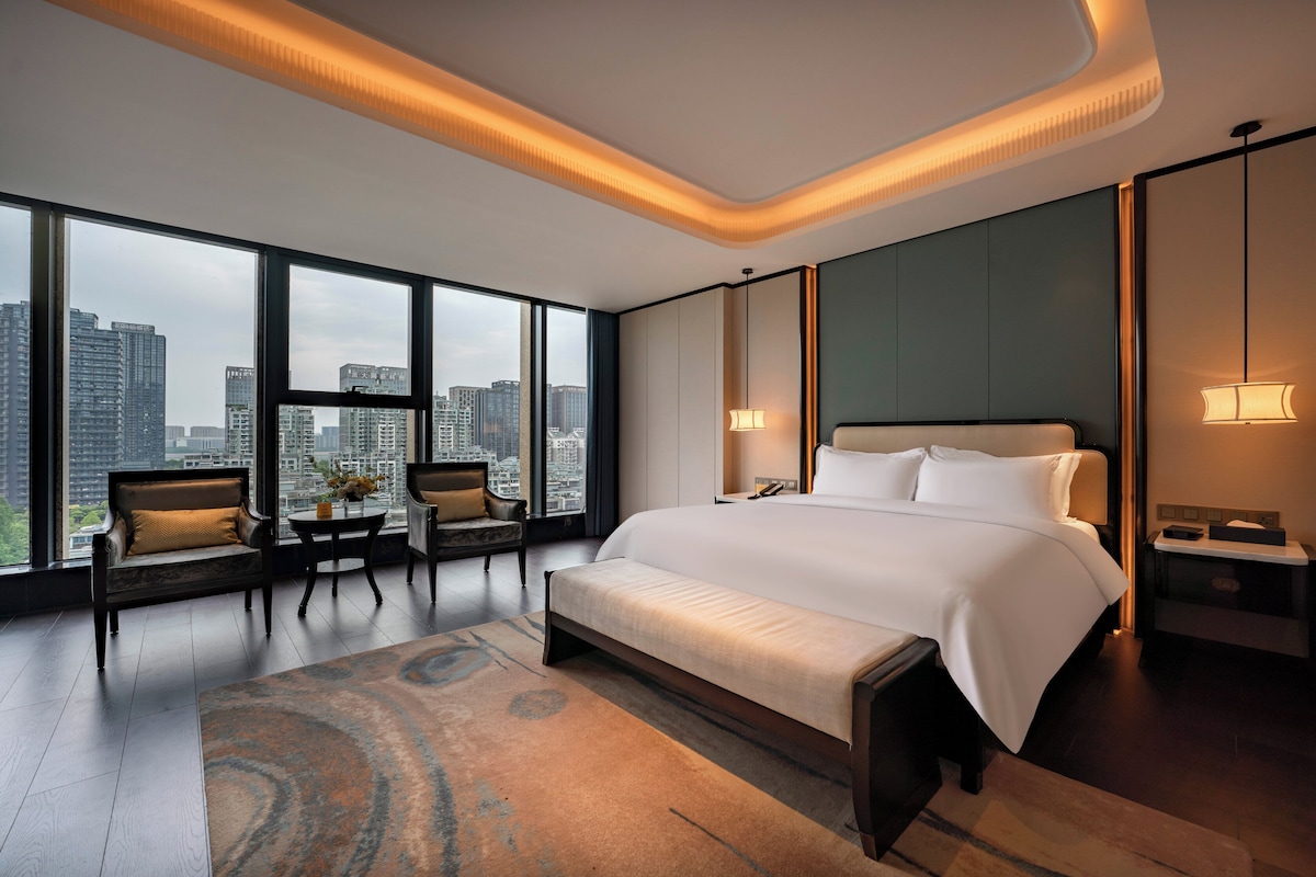 A modern hotel room with a large bed, a seating area with chairs, a view of the city, and a cityscape through the windows.