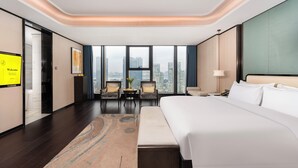 A modern hotel room with a large bed, a seating area with two chairs, a small table, and a city view through large windows. - voco Hangzhou Binjiang Minghao by IHG (Hangzhou)