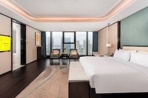 A modern hotel room with a large bed, a seating area with two chairs, a small table, and a city view through large windows.