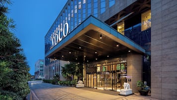 A hotel entrance with a modern design, featuring a prominent 'Voco' sign, a covered entrance, and a statue of an elephant.