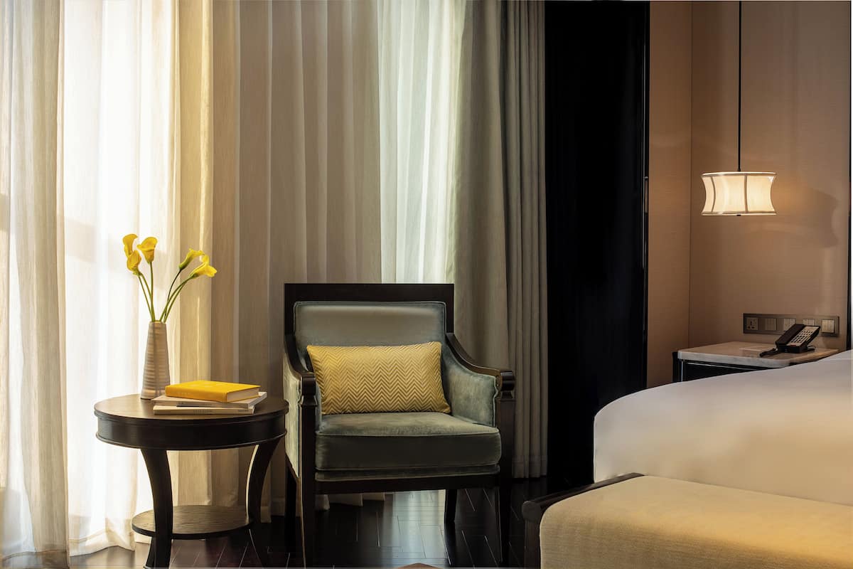 Une chambre d’hôtel avec un lit, une chaise, une petite table sur laquelle se trouvent un vase et des livres, et un téléphone sur la table de chevet.