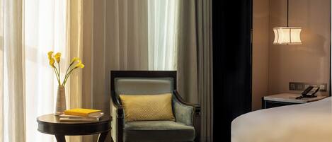 A hotel room with a bed, a chair, a small table with a vase and books, and a bedside telephone.
