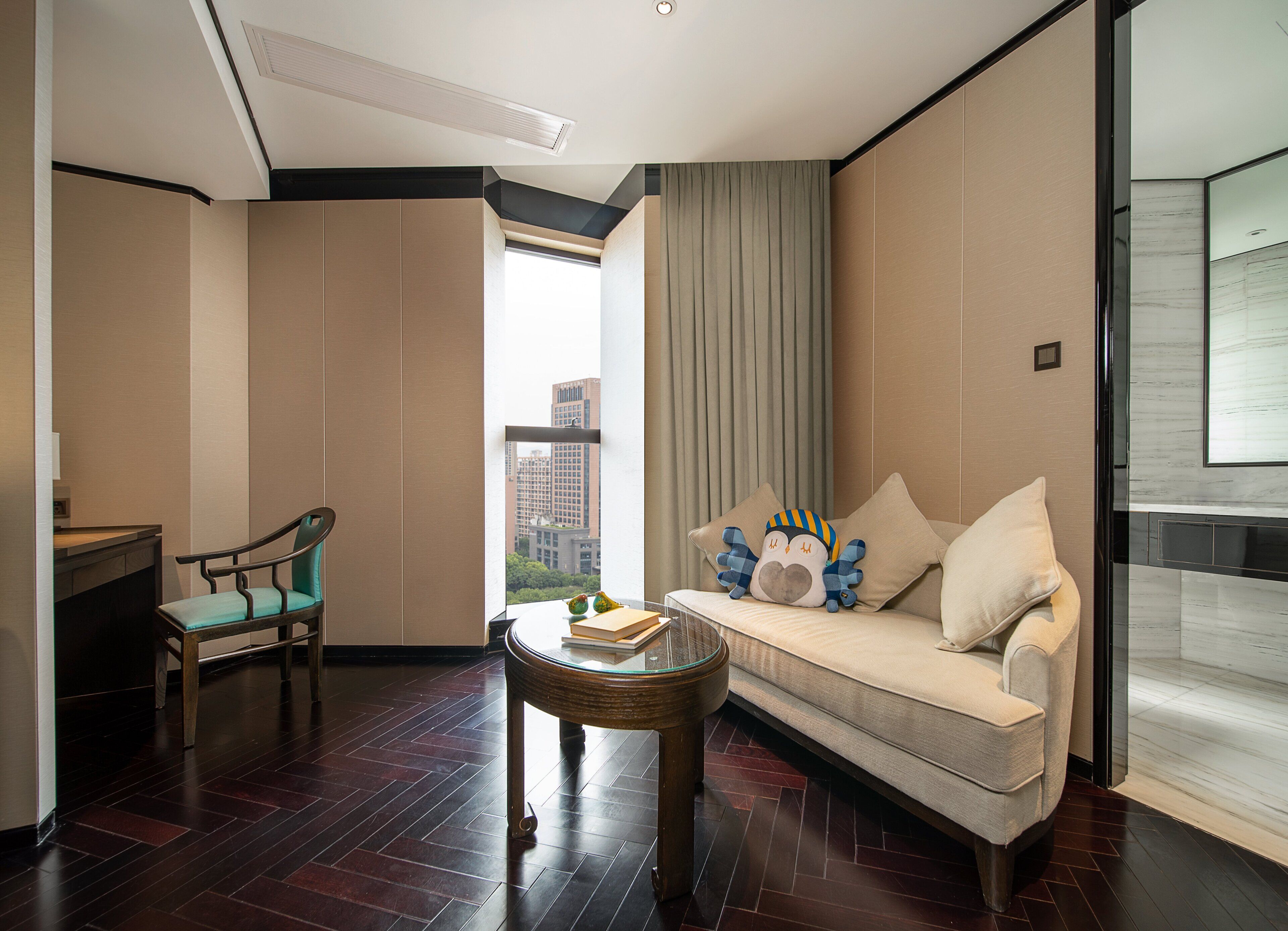 Habitación de hotel moderna con sofá, una silla, una mesita y vistas de la ciudad a través de un ventanal grande.