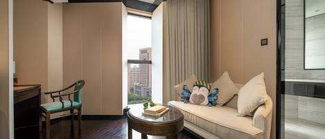 Habitación de hotel moderna con sofá, una silla, una mesita y vistas de la ciudad a través de un ventanal grande.
