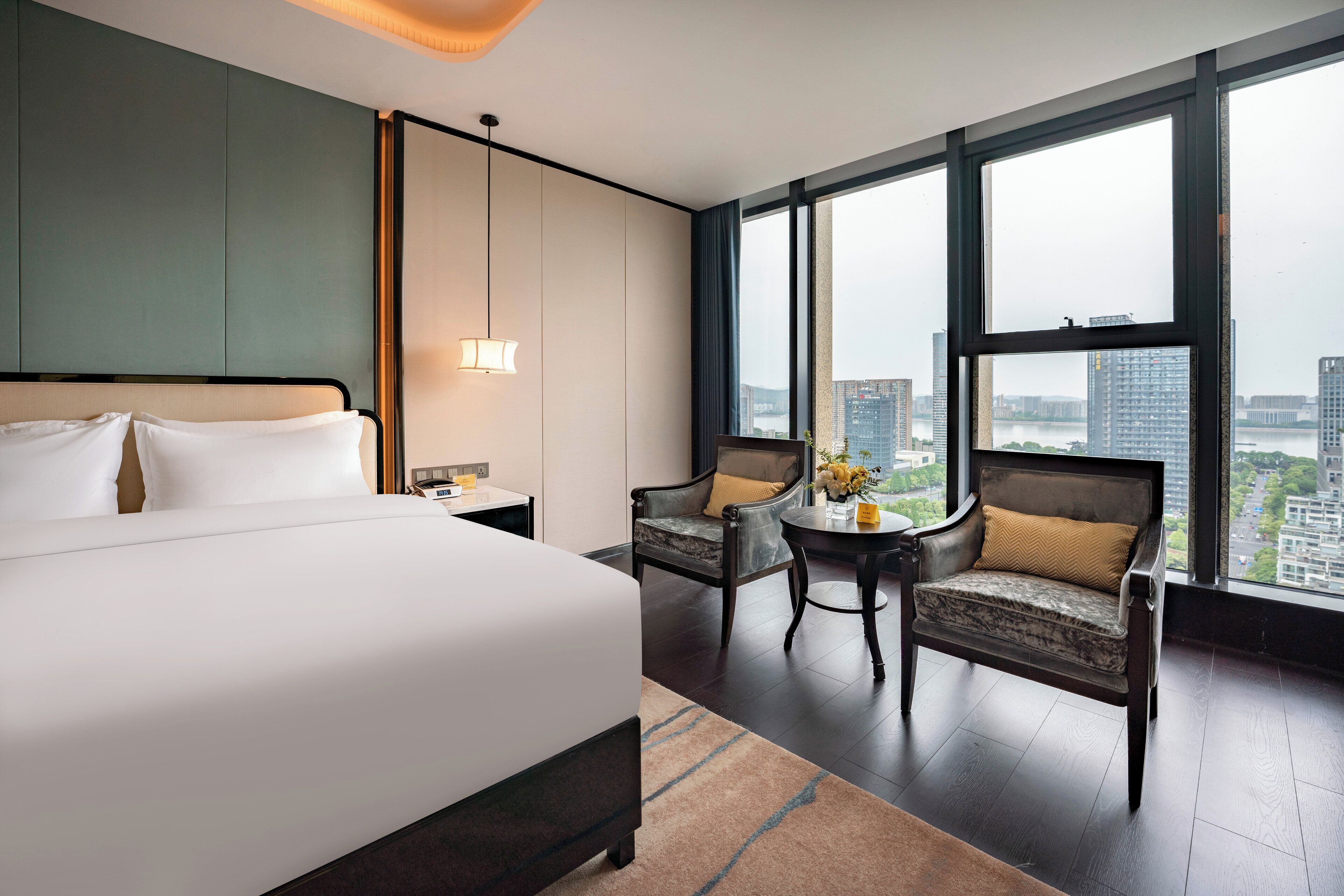 A hotel room with a large bed, a chair, a small table with flowers, and a view of the city through large windows.