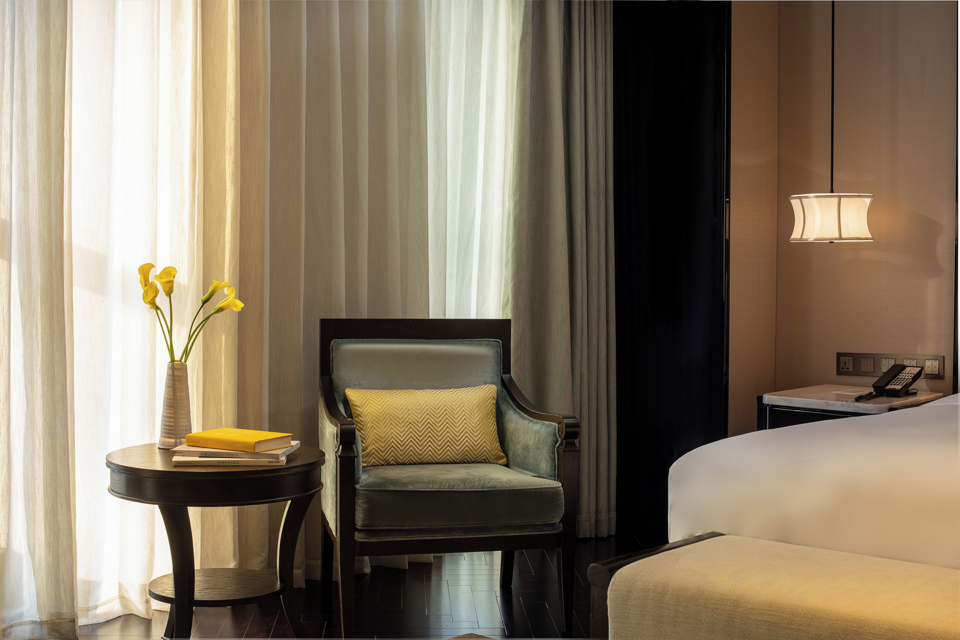 A hotel room with a bed, a chair, a small table with a vase of flowers, and a lamp.