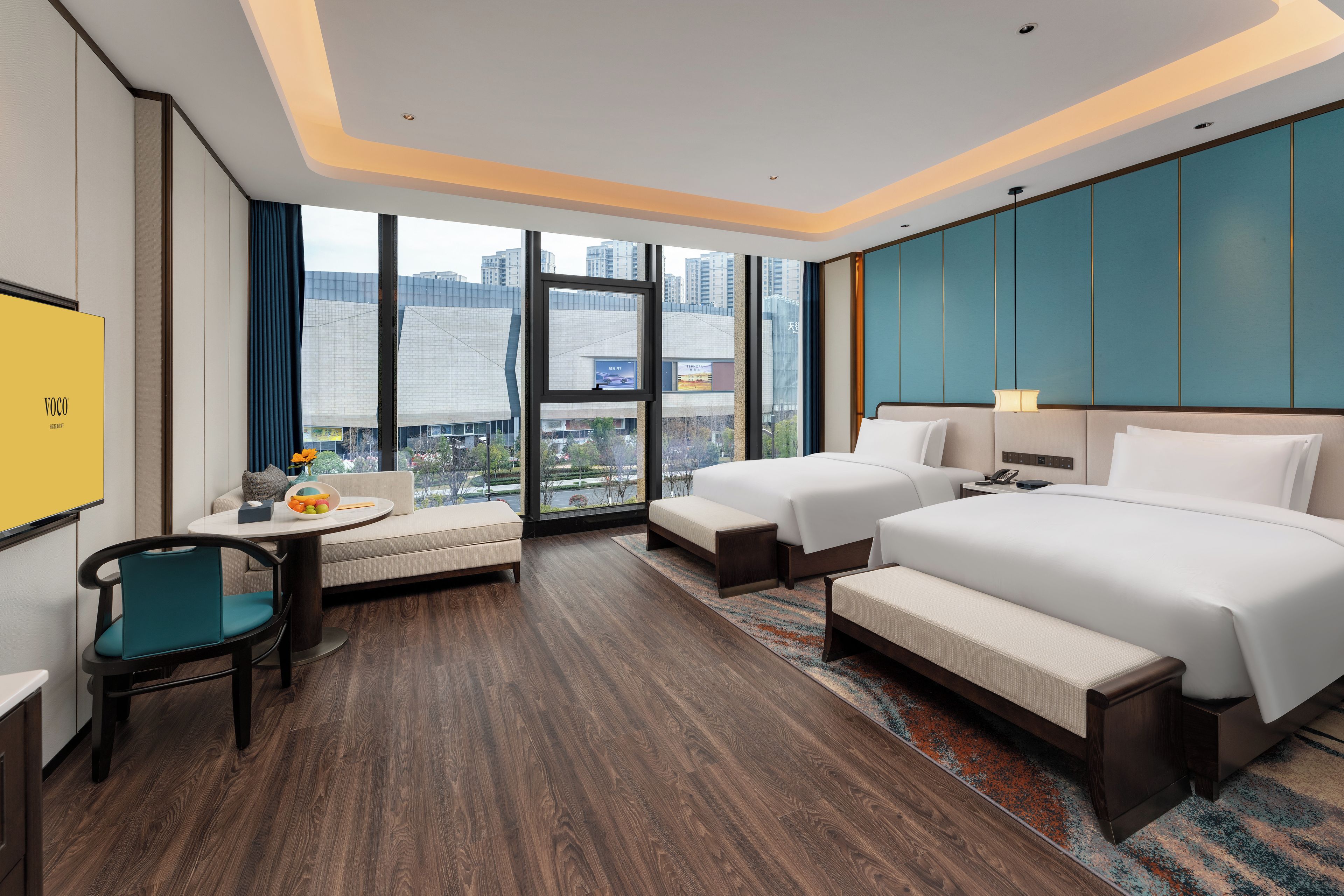 Ein Hotelzimmer mit einem großen Bett, einem kleineren Bett, einem Sofa, einem Schreibtisch und einem Stuhl. Ein Fenster bietet Blick auf Gebäude.