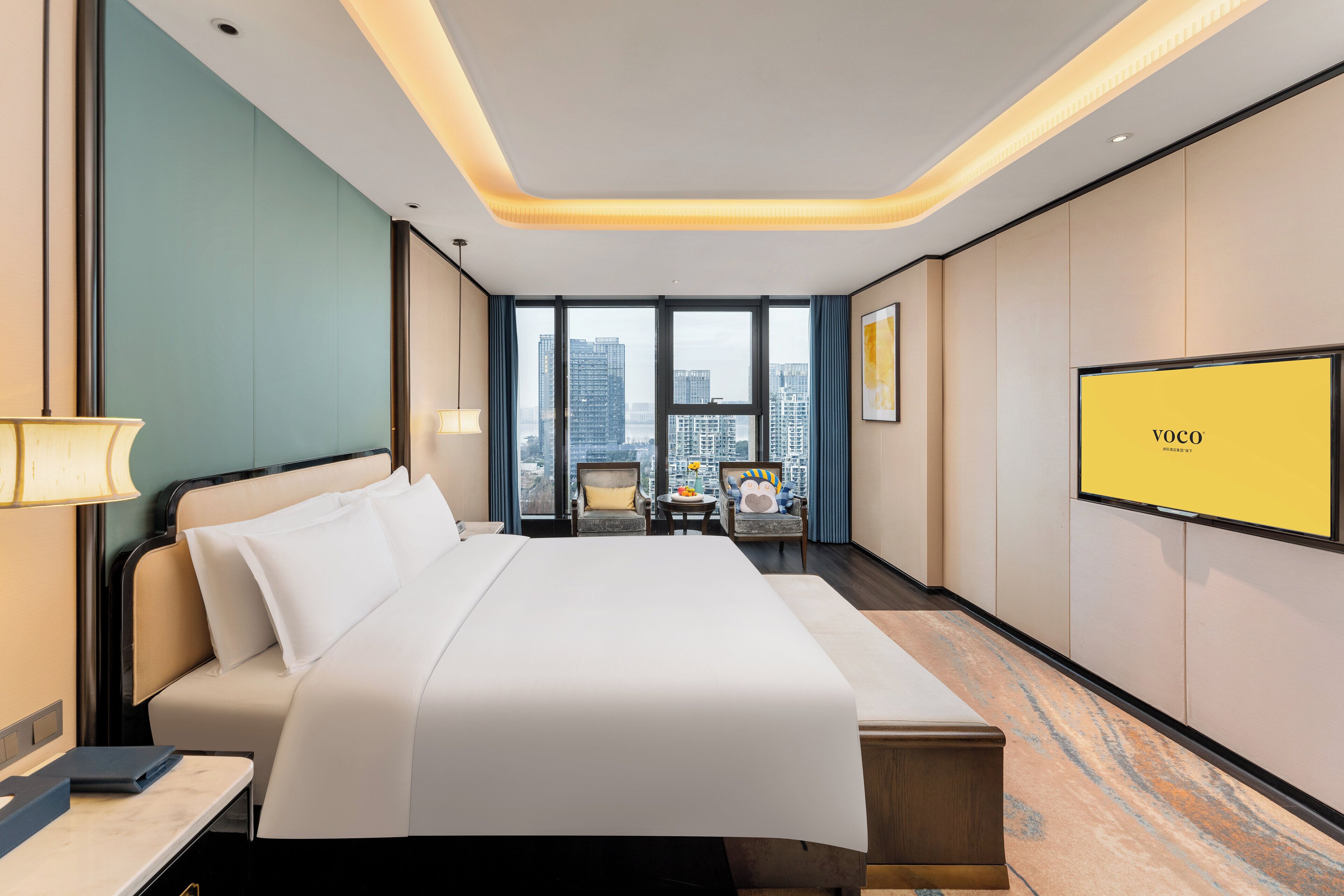 A hotel room with a large bed, a desk, a chair, a TV, and a view of the city through large windows.