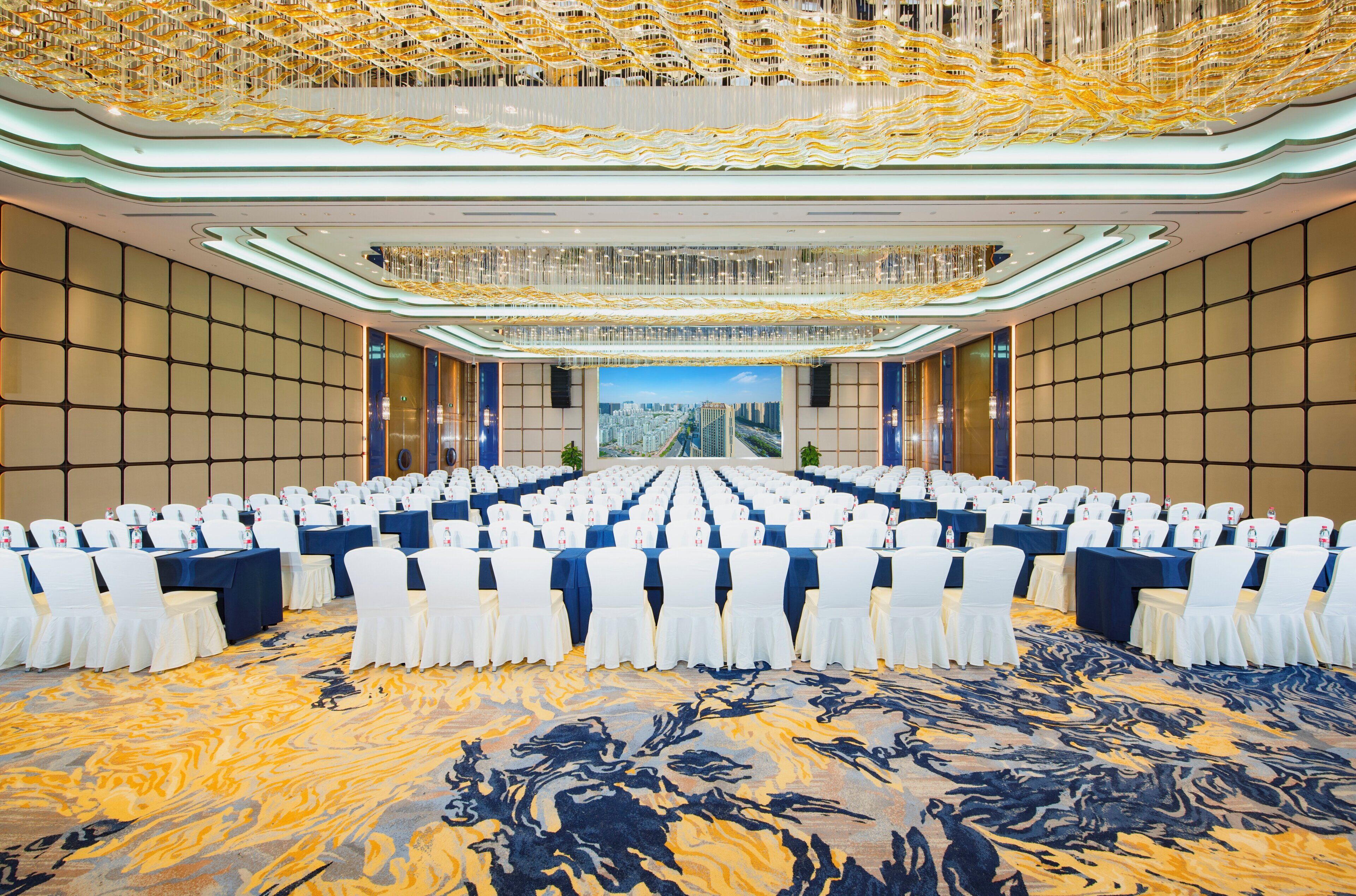 A large conference room with rows of tables and chairs, a patterned carpet, and a view of a cityscape through a large window.