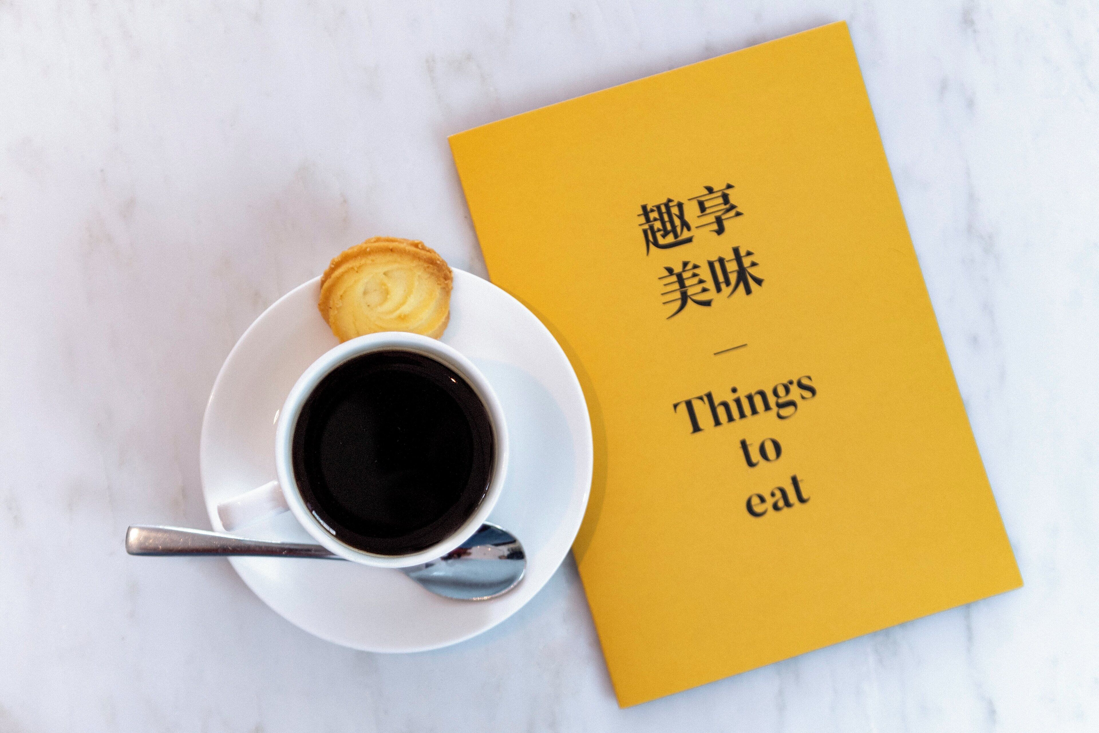 A cup of coffee on a saucer with a spoon, next to a yellow book titled 'Things to eat'.