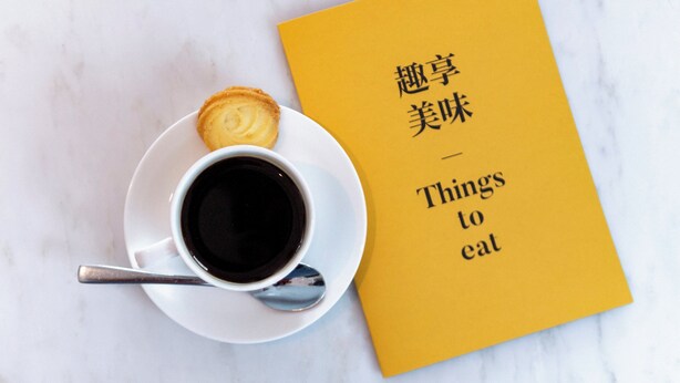 A cup of coffee on a saucer with a spoon, next to a yellow book titled 'Things to eat'. - voco Hangzhou Binjiang Minghao by IHG (Hangzhou)