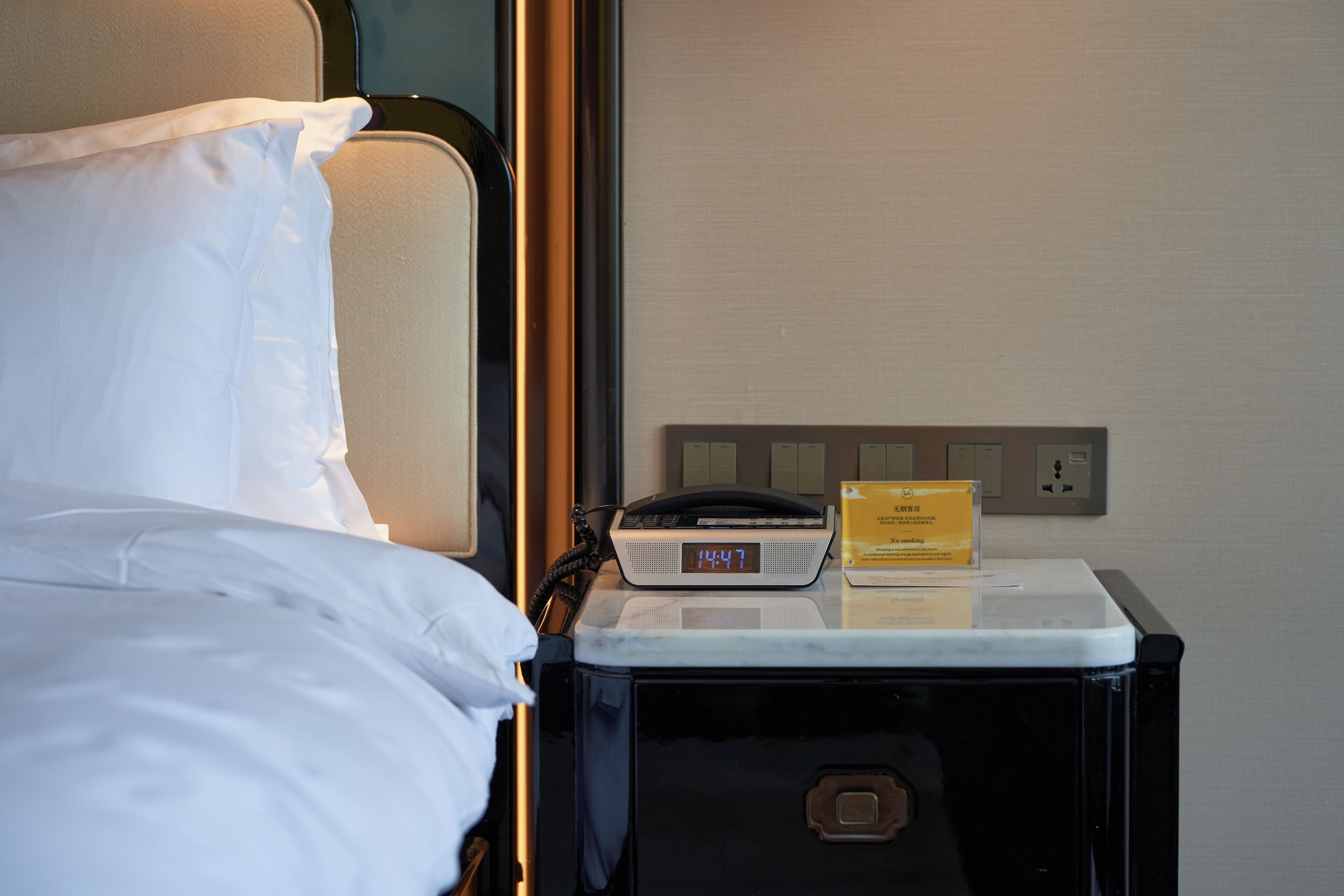 A hotel room with a bed, a nightstand, a digital clock, and a power outlet.
