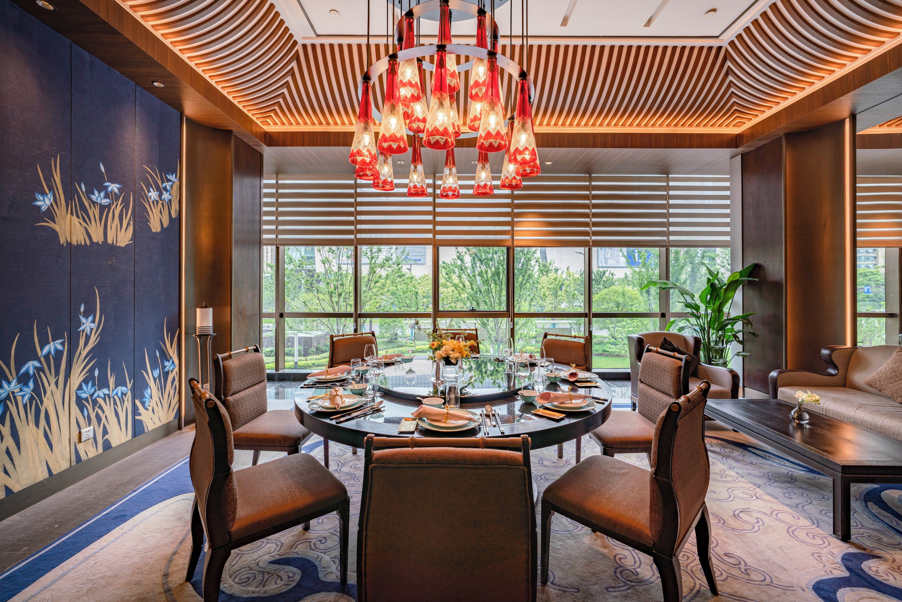 A dining area with a round table set for six, surrounded by six chairs, with a large window providing a view of greenery outside.