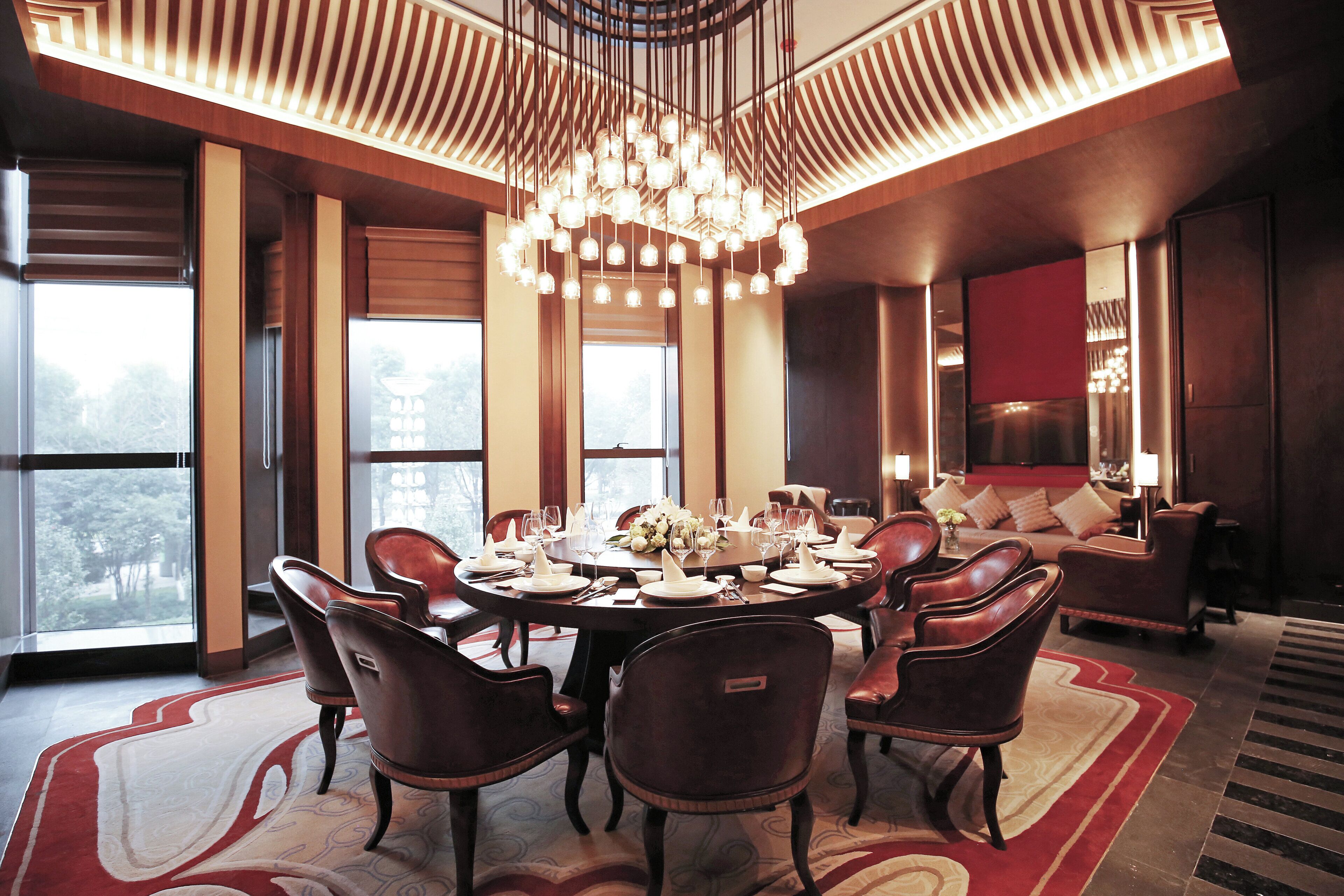 A dining area with a round table set for six, surrounded by leather chairs, with a chandelier overhead and large windows providing natural light.