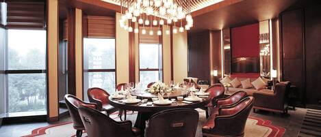 A dining area with a round table set for six, surrounded by leather chairs, with a chandelier overhead and large windows providing natural light.