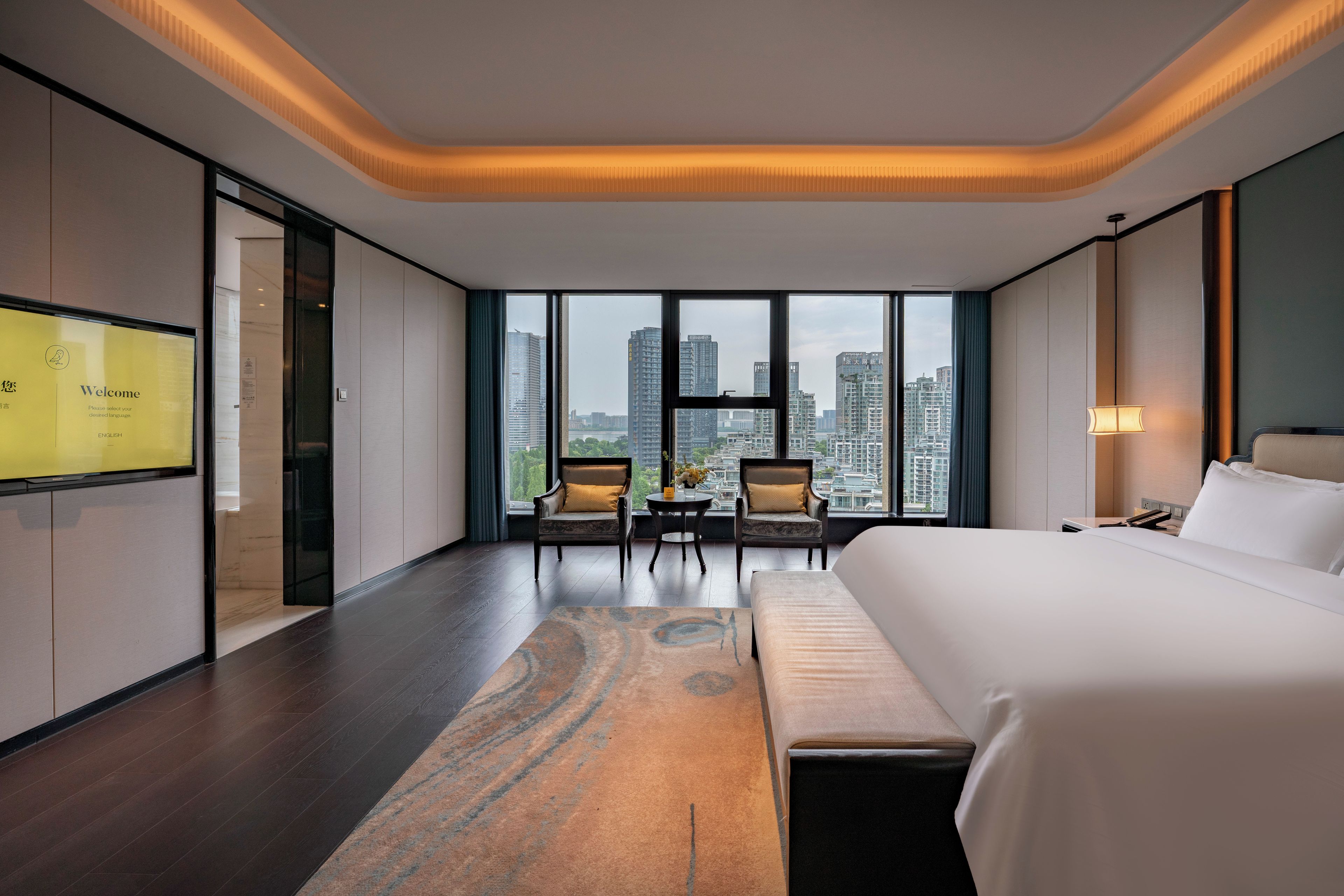 A hotel room with a large bed, a desk, two chairs, a view of the cityscape, and a television on the wall.