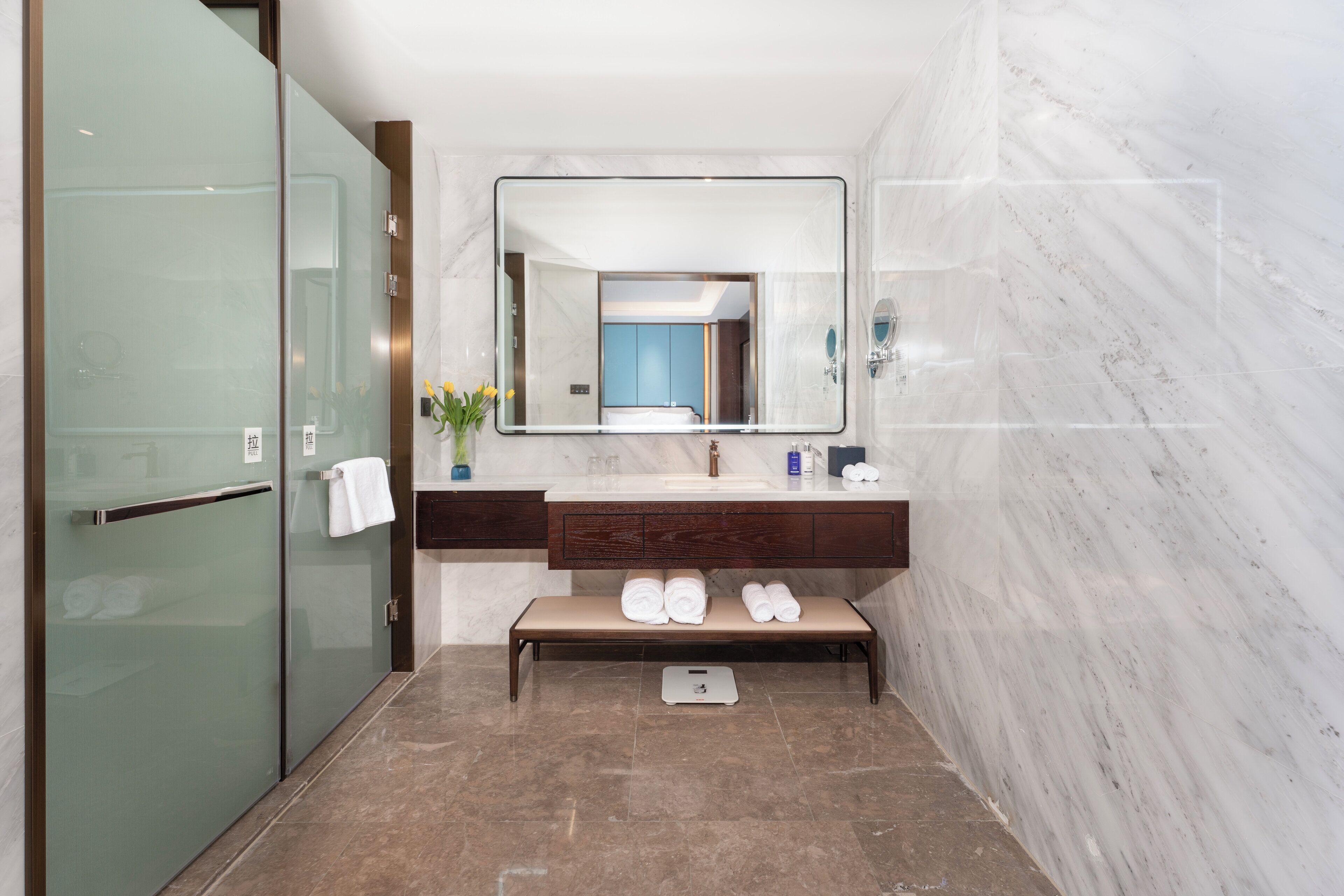 A modern bathroom with a marble wall, a large mirror, a wooden bench, and a scale.